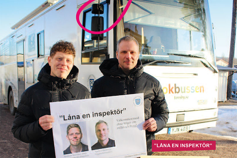 Två män framför en buss, i handen håller de en skylt med texten "Låna en inspektör"